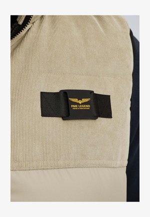 Tissu en velours côtelé beige avec une sangle noire et un patch rectangulaire noir arborant un logo d'ailes jaunes et le texte "PME Legend Tested at High Altitude".