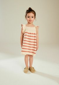 Strikkjole med bølget kant, cremefarvet med rustfarvede striber, udstyret med blonderdetaljer øverst og flagrende ærmer. Tan farvede klipklapper fuldender outfittet.