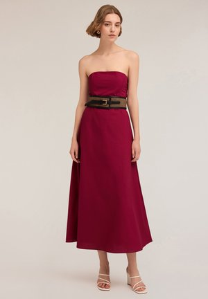 Strapless, lange jurk van diep bordeauxrood stof, met een gestructureerde zwarte riem in de taille en witte hakken.