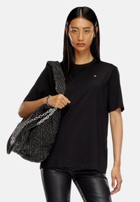 T-shirt en coton noir à manches courtes, avec un petit logo sur la poitrine ; le modèle tient un grand sac matelassé avec des accents en chaîne argentée.