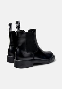 Une paire de bottines noires en cuir avec des panneaux élastiques sur les côtés, des tirettes de marque, et des talons bas nervurés, sur un fond blanc.