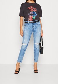 T-shirt grafica nera con un design colorato dei Rolling Stones, abbinata a jeans chiari strappati e sandali neri con tacco.