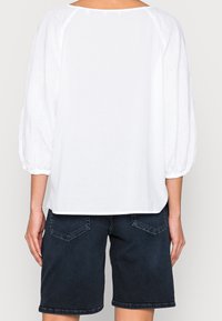 Blouse blanche à manches longues avec une coupe décontractée, des poignets froncés et un col rond, associée à un short en denim foncé avec des poches arrière.