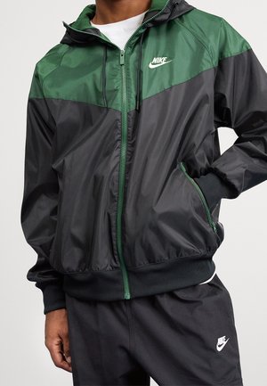 Homme portant une veste coupe-vent Nike noire et vert foncé avec des poches zippées et un pantalon Nike noir assorti.