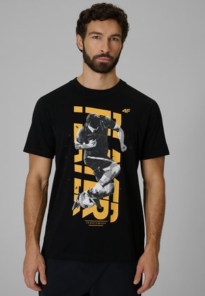 Hombre con barba que lleva una camiseta negra con un dibujo de un futbolista pateando un balón sobre un fondo geométrico amarillo.