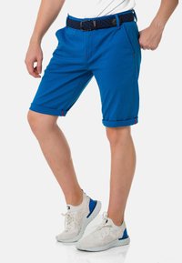 Blaue Shorts aus leichtem Stoff, mit umgeschlagenen Säumen, seitlichen Taschen und roten Nähakzenten. Kombiniert mit weißen und blauen Sneakern.