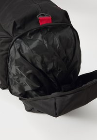 Mochila negra con el compartimento principal abierto que muestra un interior de tela negra suave y un lazo de transporte rojo en la parte superior.