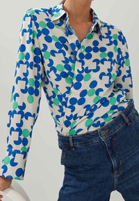 Femme portant une chemise blanche boutonnée avec des cercles géométriques bleus et verts rentrée dans un jean bleu taille haute.