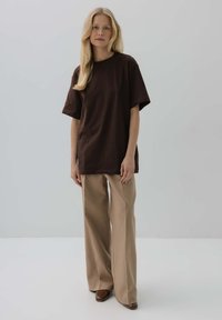Brązowa koszulka bawełniana z krótkimi rękawami, luźny krój, zestawiona z beżowymi spodniami typu wide-leg i brązowymi butami, na jednolitym tle.