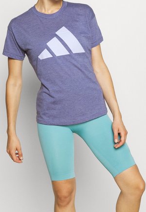 Personne portant un t-shirt violet avec des bandes triangulaires blanches et un short de cycliste ajusté bleu clair, debout devant un fond uni.