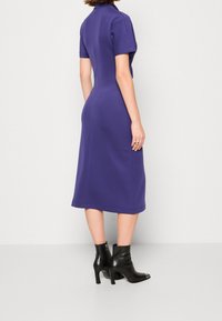 Fialové šaty do poloviny stehen s krátkými rukávy, vypasovaným pasem a rovný střih. Nosené s černými vysokými kotníkovými botami.