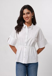 Blusa bianca a maniche corte con bottone frontale e vita elastica. Realizzata in tessuto strutturato, presenta un colletto e dettagli di maniche plissettate.