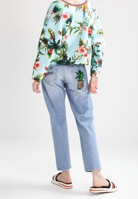 Blusa de manga larga tropical de color azul claro con estampado floral, combinada con jeans de mezclilla azul claro que presentan un diseño de piña con lentejuelas en el bolsillo trasero.