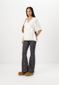 Witte eyelet wikkeltop met korte mouwen, gecombineerd met zwart flared pantalons met een bloemenpatroon; bruine platform schoenen en een bruine handtas.