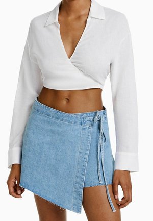 Kvinde iført en hvid langærmet cropped wrap skjorte og et asymmetrisk lyseblåt denim mini-nederdel med bindebånd i siden.