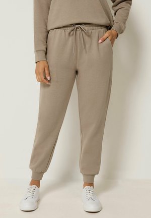 Loszittende taupekleurige joggingbroek met elastische manchetten en een taille met trekkoord, gedragen met witte sneakers, één hand in de zak.