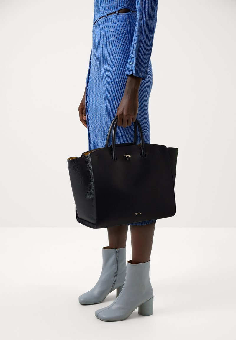 Černá kožená taška přes rameno s texturovaným povrchem a dvojitými uchy; logo zobrazené v přední části. Modelka má na sobě modré žebrované šaty a šedé kotníkové boty.