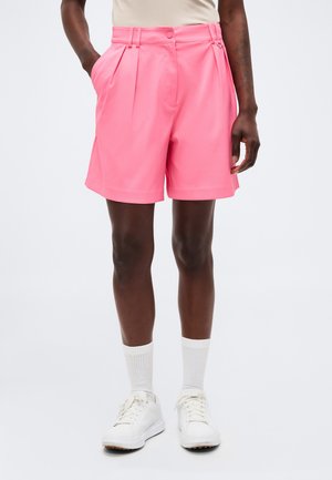 Personne à la peau noire portant un short rose vif sur mesure, des chaussettes blanches, des baskets blanches et un haut beige, debout avec une main dans la poche.