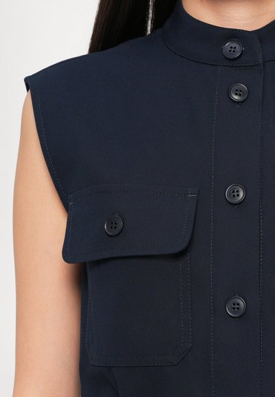 Navy sleeveless shirt with a stand collar, featuring front button closure and a patch pocket with a flap. Smooth fabric with visible stitching.