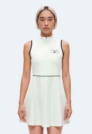 Femme aux cheveux blonds courts portant une robe blanche sans manches avec un col zippé et une bordure noire, debout devant un fond uni.