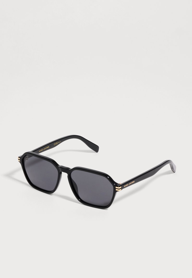 Lunettes de soleil rectangulaires noires avec verres foncés et accents dorés aux coins de la monture et sur les branches, sur un fond clair.