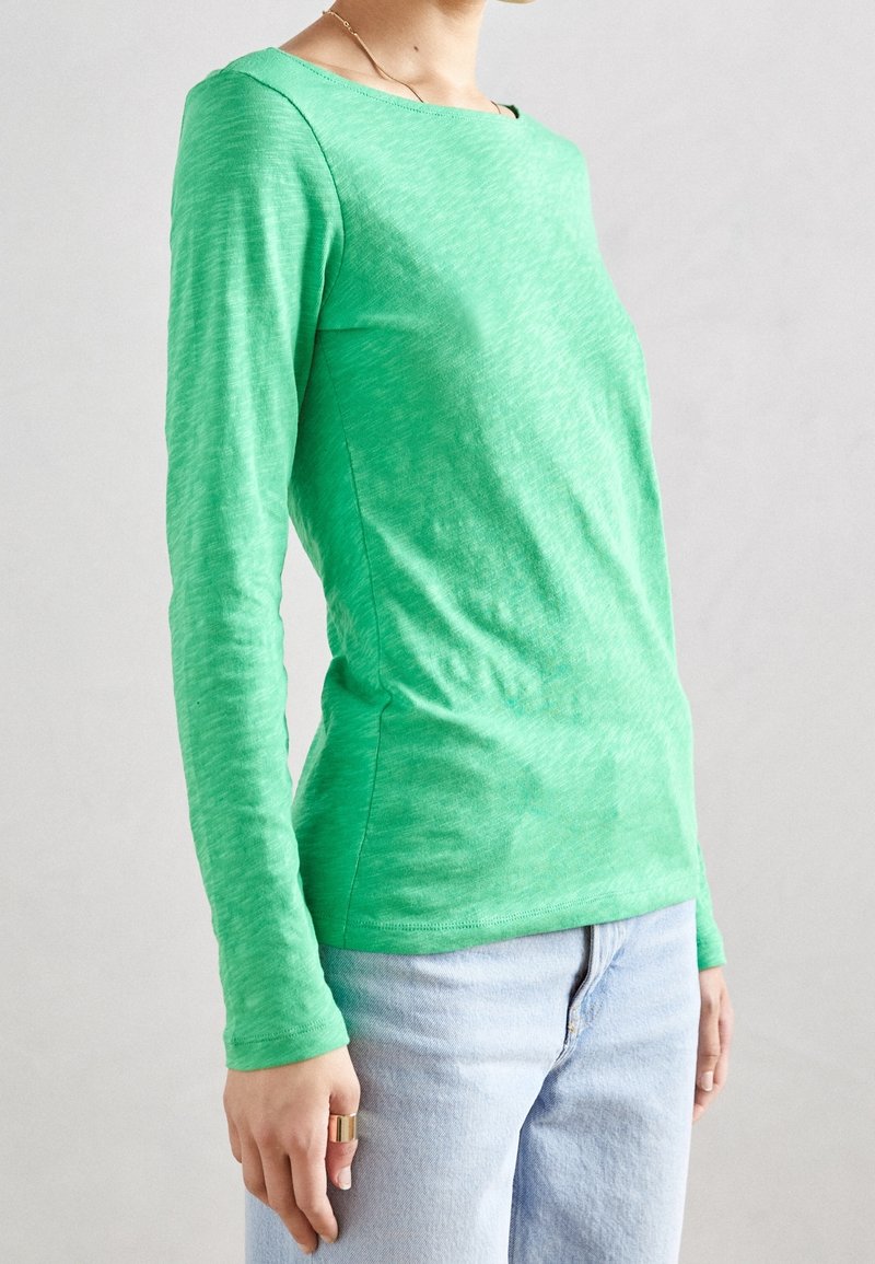 Maglietta a maniche lunghe di colore verde realizzata in tessuto leggero e testurizzato, con scollatura a barca e silhouette aderente, abbinata a jeans in denim chiaro.