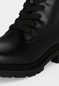 Gros plan d'une botte en cuir noir à lacets avec une semelle épaisse crantée et un petit talon bloc sur fond blanc.