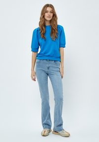 Blå kortärmad sweatshirt med ribbad muddar, kombinerad med ljusblå utsvängda jeans. Skorna är beige med orange detaljer. Enkel bakgrund.