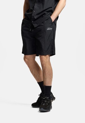 Mężczyzna ubrany w czarne szorty z białym logo, czarną koszulę na guziki, czarne skarpety i czarne buty sportowe, stoi z rękami w kieszeniach.