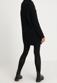 Schwarzes Strickkleid mit gerippter Textur, das bis zur Mitte des Oberschenkels reicht, kombiniert mit schwarzen Leggings und Stiefeletten. Hat ein kragenförmiges Design.