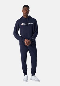 Marineblauw trainingspak van zacht materiaal, met een hoodie met een voorzak en een wit Champion-logo; gecombineerd met bijpassende joggingbroek.