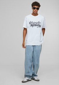 Hvid grafisk T-shirt med "NEW YORK" i sort tekst, parret med løse lyseblå jeans og sort-hvide sneakers. Model iført solbriller.