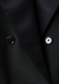 Chaqueta negra con tela texturizada, que presenta dos botones: uno oscuro y brillante y un botón redondo blanco con un diseño negro.