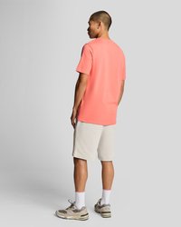 Koraļļu krāsas īsām piedurknēm T-krekls ar brīvu piegriezumu, saskaņots ar gaišām šortiem. Modelis valkā bēša krāsas sporta apavus ar baltām zeķēm. Skats no aizmugures.