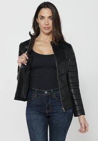 Veste en cuir noir avec des panneaux matelassés horizontaux, fermeture éclair et col à pression, portée sur un débardeur noir, associée à un jean bleu foncé.