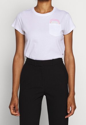 Kobieta ubrana w biały t-shirt z krótkim rękawem z małym kolorowym wzorem na kieszeni oraz czarne spodnie z wysokim stanem na jednolitym tle.