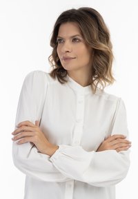 Biała koszula z guzikami o mandaryńskim kołnierzyku, puffowanych rękawach i elastycznych mankietach, wykonana z lekkiego materiału. Gładka tekstura, bez wzorów.