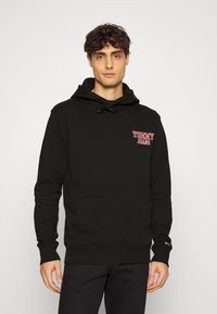 Sudadera negra de algodón, con bolsillo canguro, capucha con cordón y logotipo rojo de "TOMMY JEANS" en el pecho. Puños acanalados.