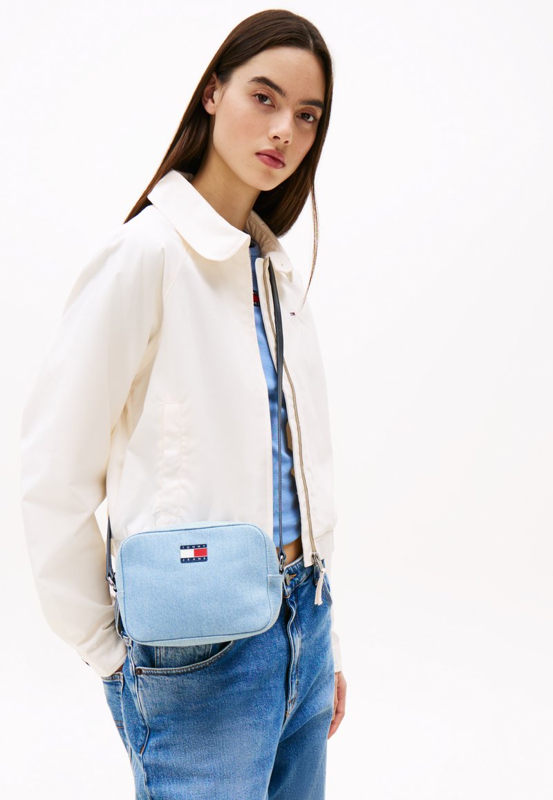 Bolso bandolera de denim azul claro con cierre de cremallera, forma rectangular y un pequeño parche con logo. El modelo lleva puesto una chaqueta blanca y una blusa azul.