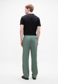 Schwarzes kurzärmliges Shirt, kombiniert mit hellgrünen gemusterten Hosen mit einer Gesäßtasche und lockerer Passform. Das Modell wird von hinten gezeigt.