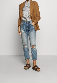 Blazer marrón sobre una blusa azul con flores y volantes, combinado con jeans claros desgastados y sandalias marrones con detalles de hebilla.