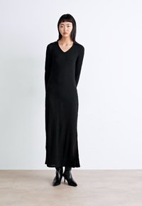 Vestido longo preto canelado com decote em V, mangas longas e uma silhueta fluida. Usado com botas de tornozelo pretas de bico fino.