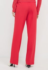 Rote, weit geschnittene Hosen aus glattem Stoff mit einem taillierten Schnitt und einer Mittelfalte. Kombiniert mit schwarzen Heels für einen schlanken Look.