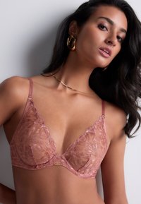 Soutien-gorge à décolleté plongeant avec dentelle en rose poussiéreux, orné d'un motif floral complexe et de panneaux transparents. Il présente des bretelles fines et ajustables, ainsi que des accessoires en or.