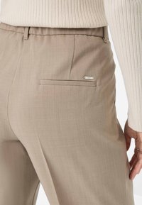 Pantalons tailleur beige avec une texture lisse, dotés d'une poche passepoilée et d'un détail logo en métal au dos.