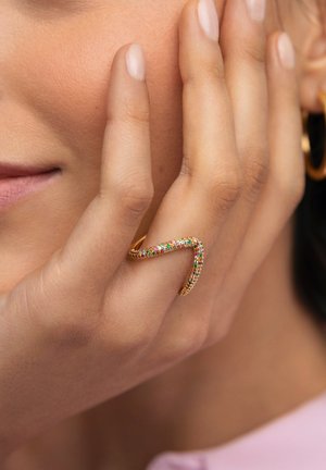 Anillo de tono dorado con un diseño ondulado, con piedras multicolores incrustadas a lo largo de la banda. Textura suave, llevado en una mano que toca el rostro.