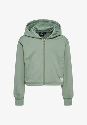 Ung pige smilende iført grøn lynlås hoodie, matchende leggings, stribede hvide sokker og blå sneakers med hvide chevron-detaljer.