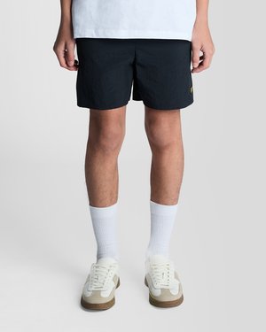 Hombre con pantalones cortos negros, calcetines blancos hasta la mitad de la espinilla y zapatillas deportivas blancas, de pie contra un fondo liso gris claro.