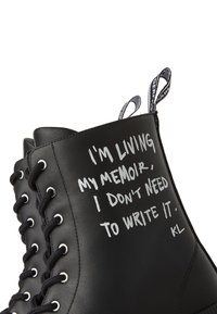 Čierne kožené členkové čižmy s bielym textom, s kovovými očkami, čiernymi šnúrkami a kontrastnými pútkami na pätách.