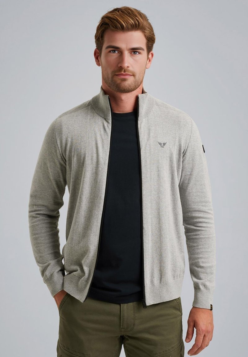 Hellgraue Zip-Jacke mit Stehkragen und gestrickter Textur, ausgestattet mit schwarzem Reißverschluss und Logo-Akzent auf der Brust. Wird über einem schwarzen Hemd getragen.